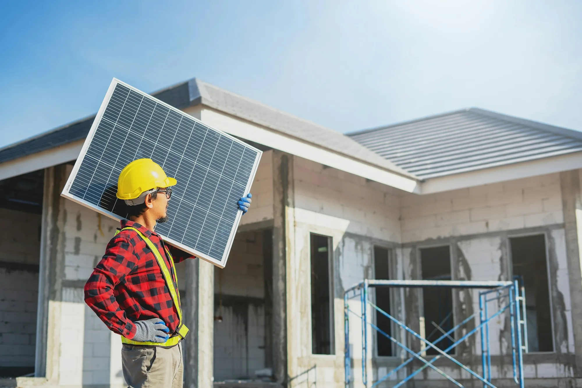 Worker in a hard hat and plaid shirt carrying a solar panel past a house under construction